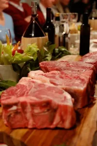 Thick raw Bistecca steaks displayed on a wooden board with fresh vegetables and wine bottles in the background at a restaurant table.