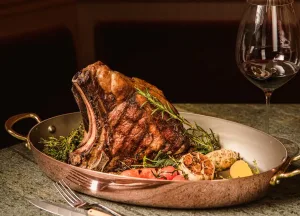 Baked steak in a pan garnished with roasted garlic and rosemary, served alongside a glass of red wine.