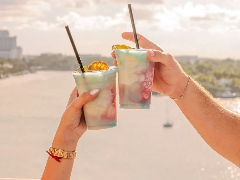 Two people toasting cocktails with a waterfront sunset view during happy hour in Miami Beach