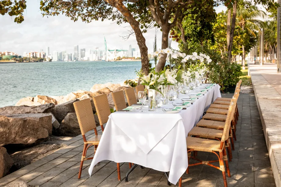Elegant outdoor waterfront dining table set with white linens and floral arrangements overlooking Biscayne Bay and the Miami skyline.