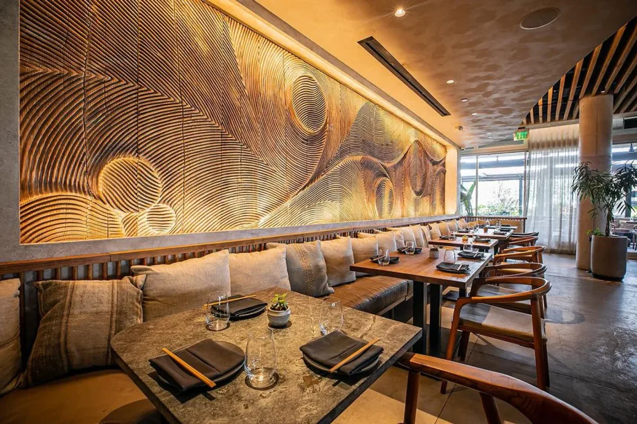Modern restaurant with warm lighting, wooden tables, cushioned bench seating, and a large textured wood wall mural.