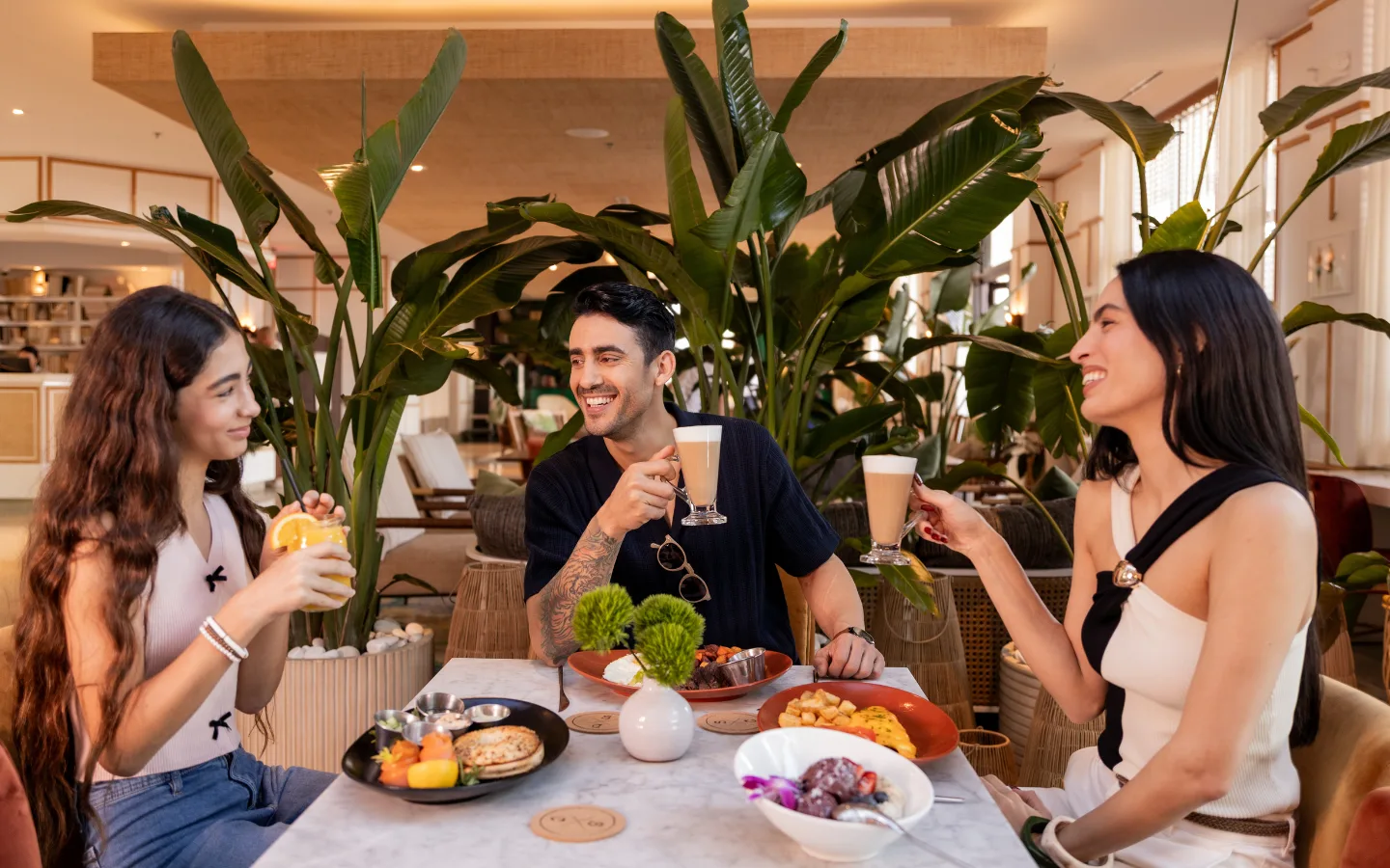 Friends enjoying Easter brunch with coffee and breakfast dishes at a stylish Miami Beach restaurant patio surrounded by tropical plants