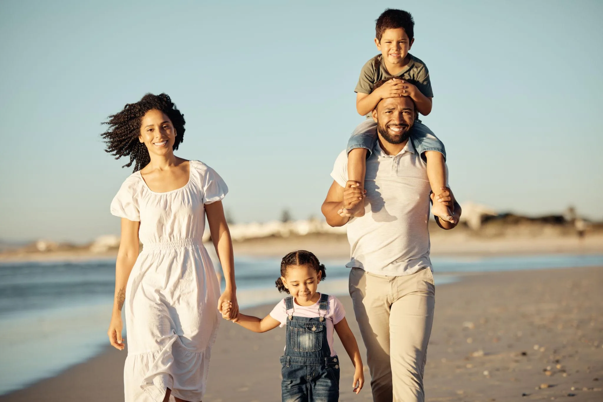 Happy family walking on the beach at sunset, father carrying child on shoulders while mother and daughter hold hands