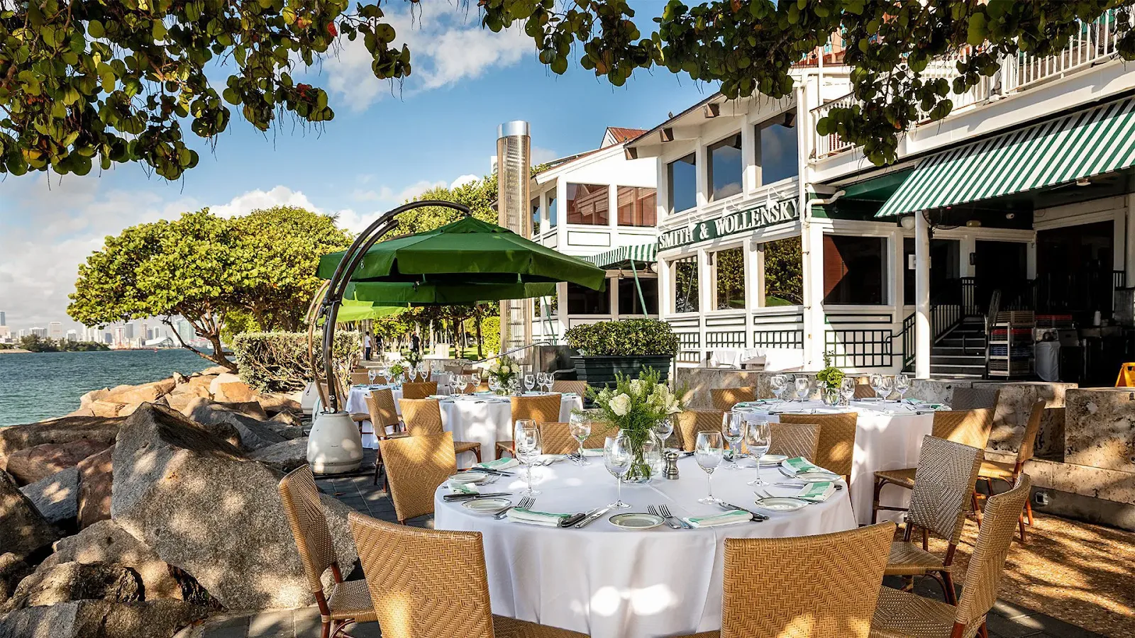 Upscale waterfront restaurant in Miami with outdoor dining tables, tropical greenery, and scenic bayfront views during daytime.