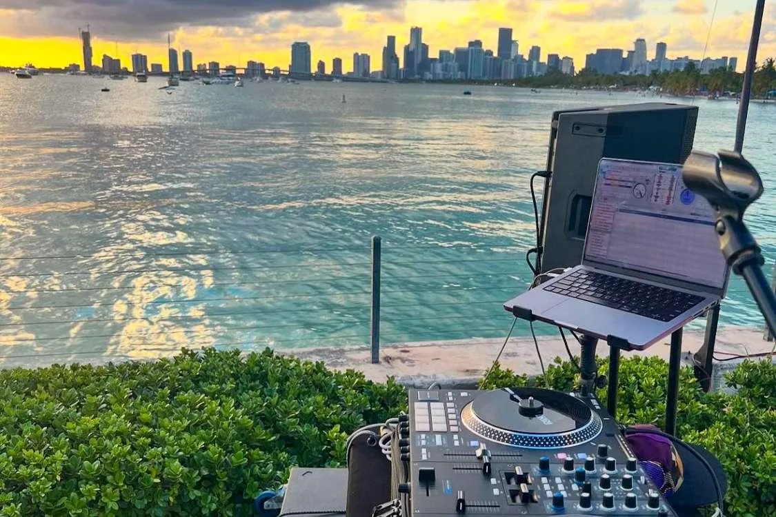 DJ setup with laptop and turntable overlooking Miami waterfront skyline at sunset, perfect for outdoor event vibes