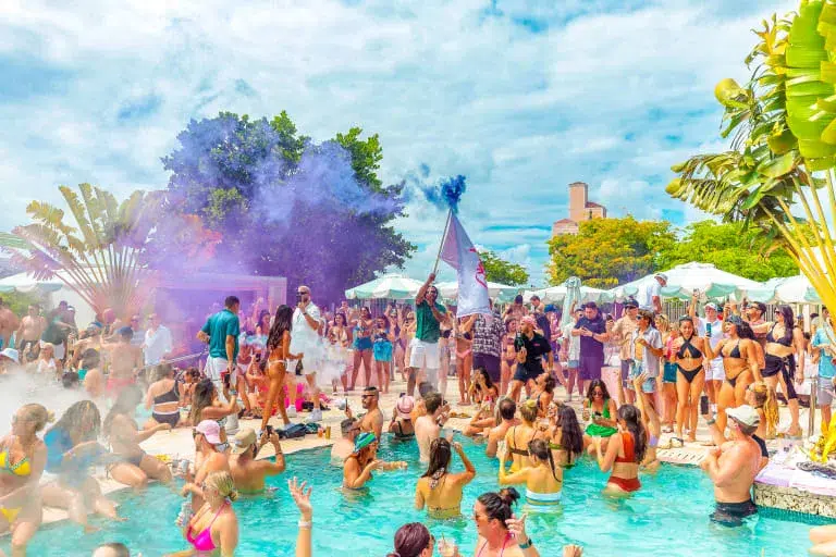 Crowded Miami pool party with DJs, colorful smoke effects, and guests celebrating during Memorial Day Weekend in Miami Beach.