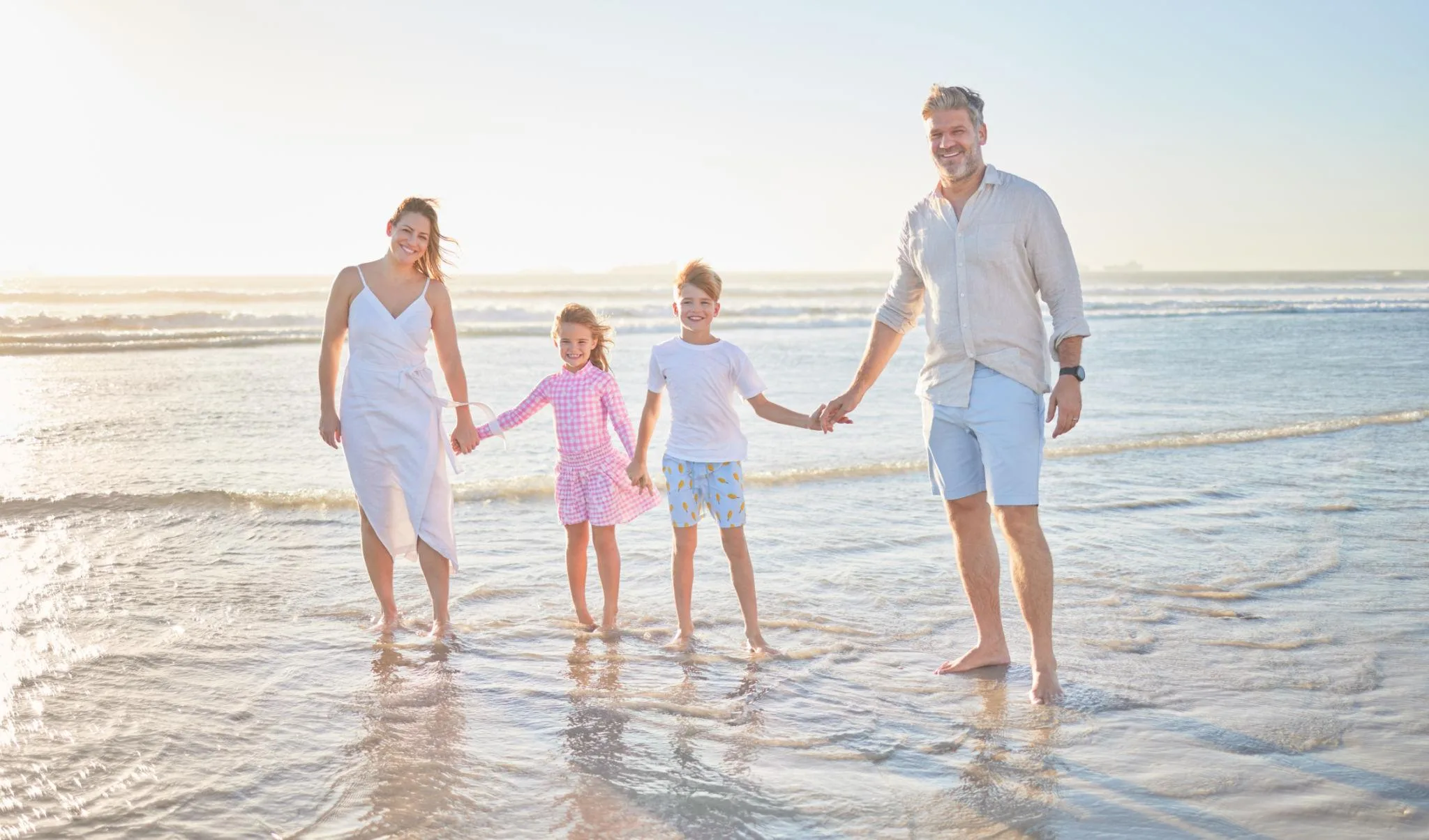 Family of four walking barefoot in the ocean on a sunny beach day, enjoying a peaceful seaside moment together