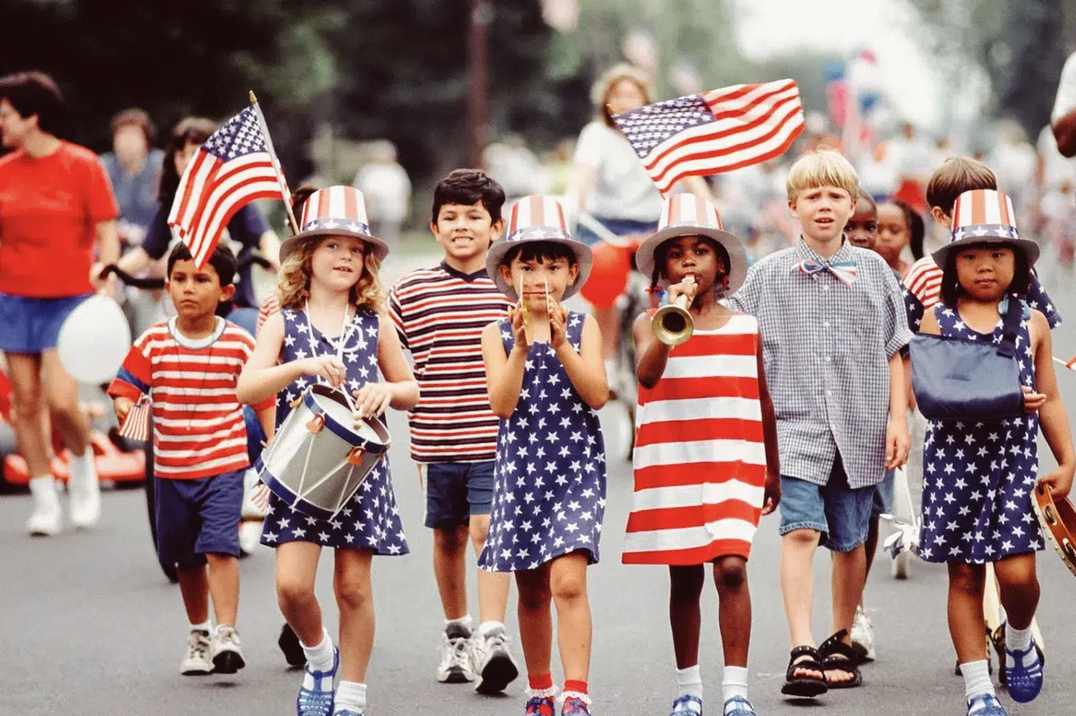 Children dressed in red, white, and blue marching in a Fourth of July parade while carrying American flags and musical instruments during a patriotic community celebration.