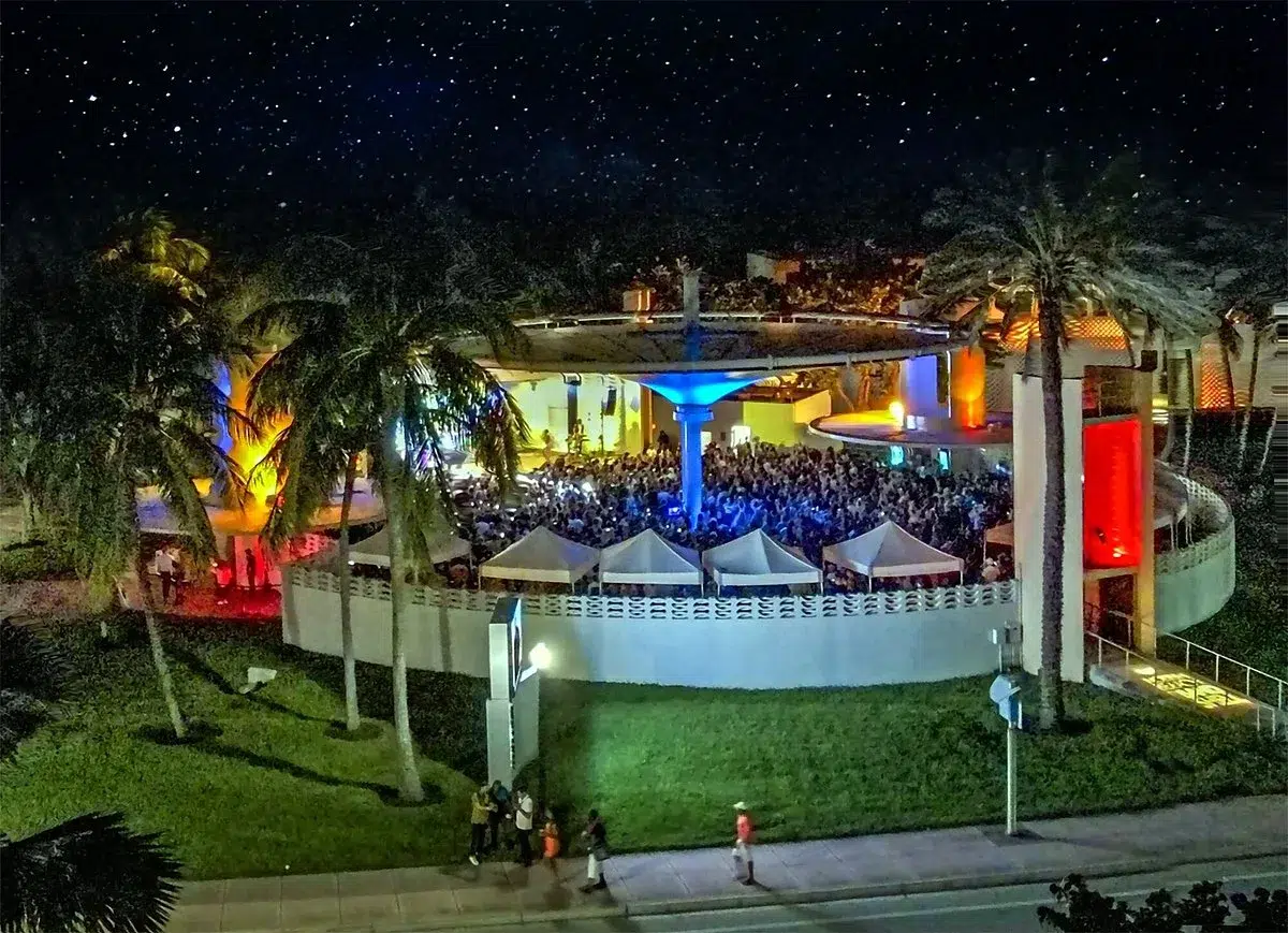 Nighttime aerial view of the Miami Beach Bandshell hosting a large outdoor concert, surrounded by palm trees and crowds attending a live entertainment event.