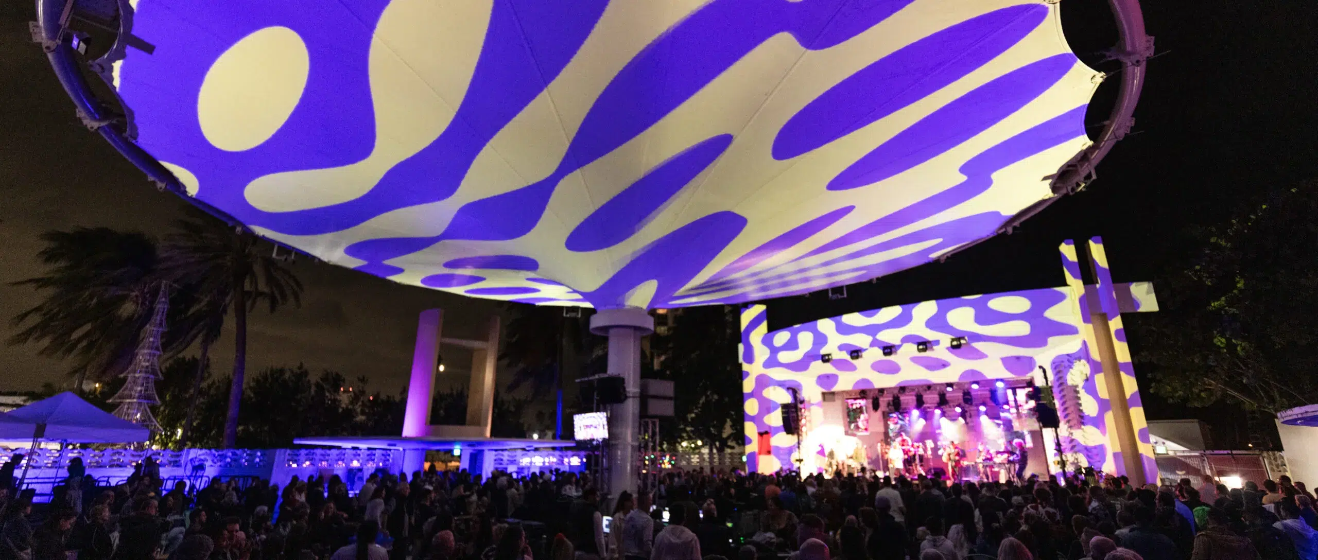Crowd gathered at the Miami Beach Bandshell during an evening concert featuring colorful stage lighting, live performances, and a vibrant outdoor festival atmosphere.