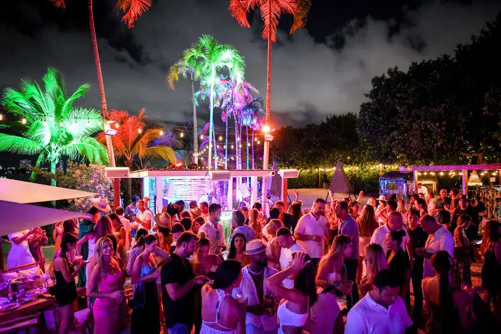 Guests enjoying an outdoor Bastille Day celebration at a Miami waterfront venue with colorful lighting, live music, palm trees, and a festive summer atmosphere.