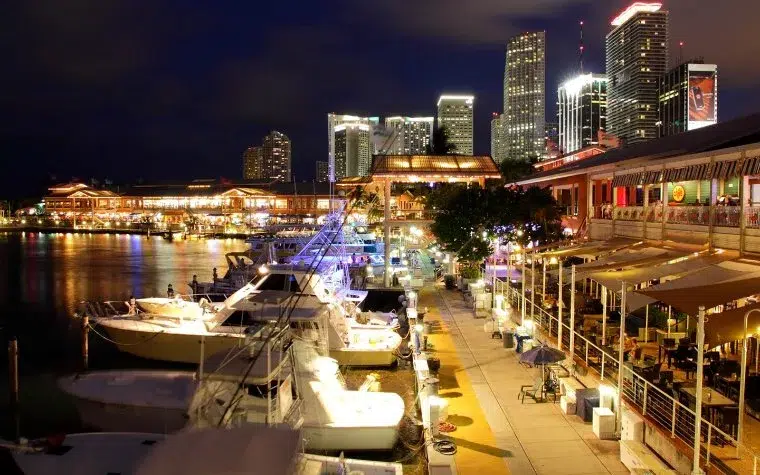 Luxury yachts docked at Bayside Marina in Downtown Miami at night, with waterfront restaurants, illuminated walkways, and the Miami skyline in the background.