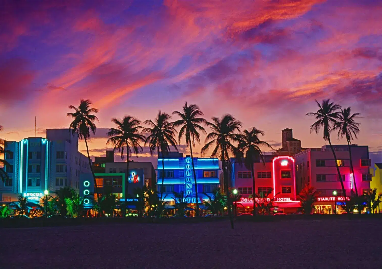 Colorful Art Deco buildings glowing along Ocean Drive in Miami Beach at sunset, framed by palm trees and a vibrant pink and purple sky.