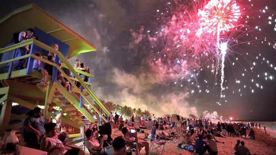 Fireworks light up the night sky above Miami Beach as crowds gather along the shoreline near a colorful lifeguard tower during a festive celebration.